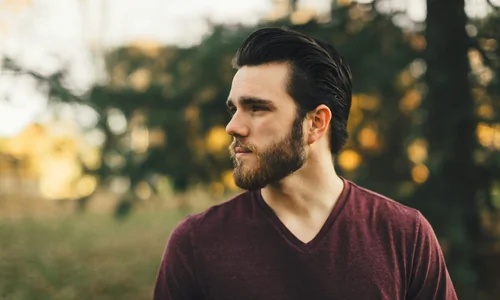 Man with a beard in a burgundy shirt, gazing in thought, representing Cognitive Behavioral Therapy (CBT).