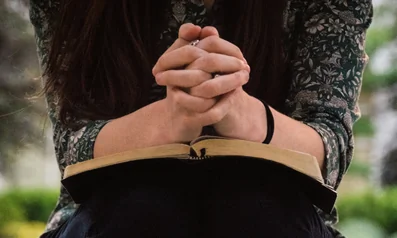 Woman with hands folded, resting on an open Bible, representing Christian counseling.